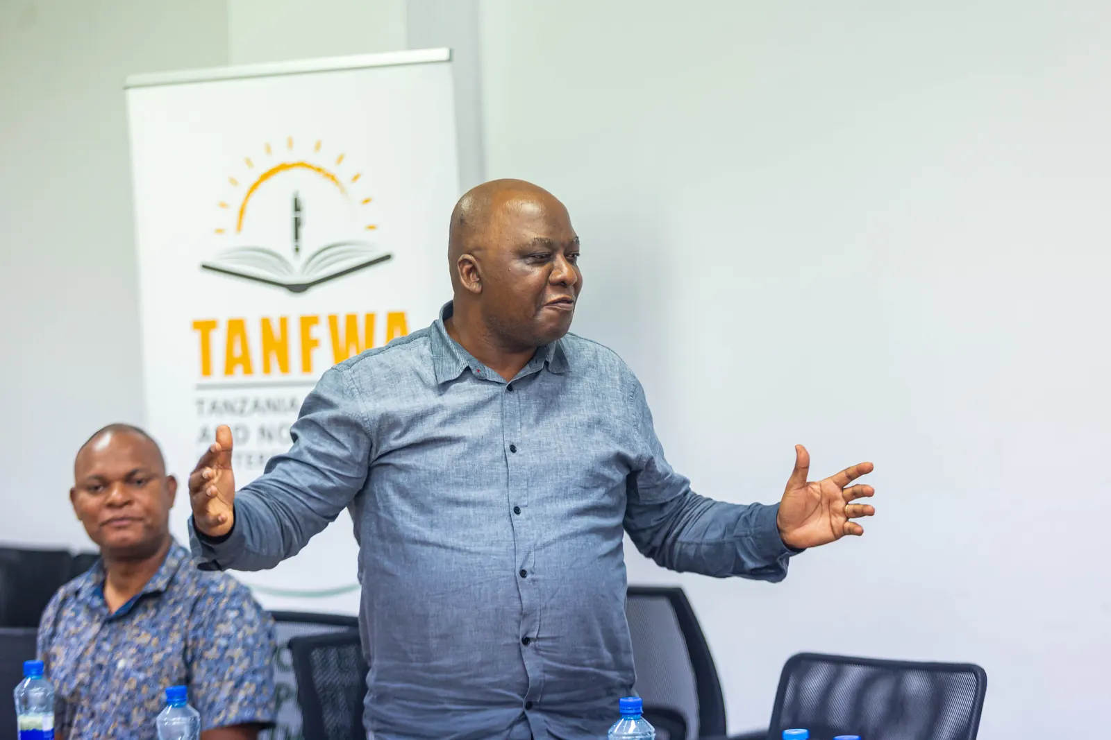 A TANFWA member speaking from the floor with his arms open, the TANFWA banner behind him